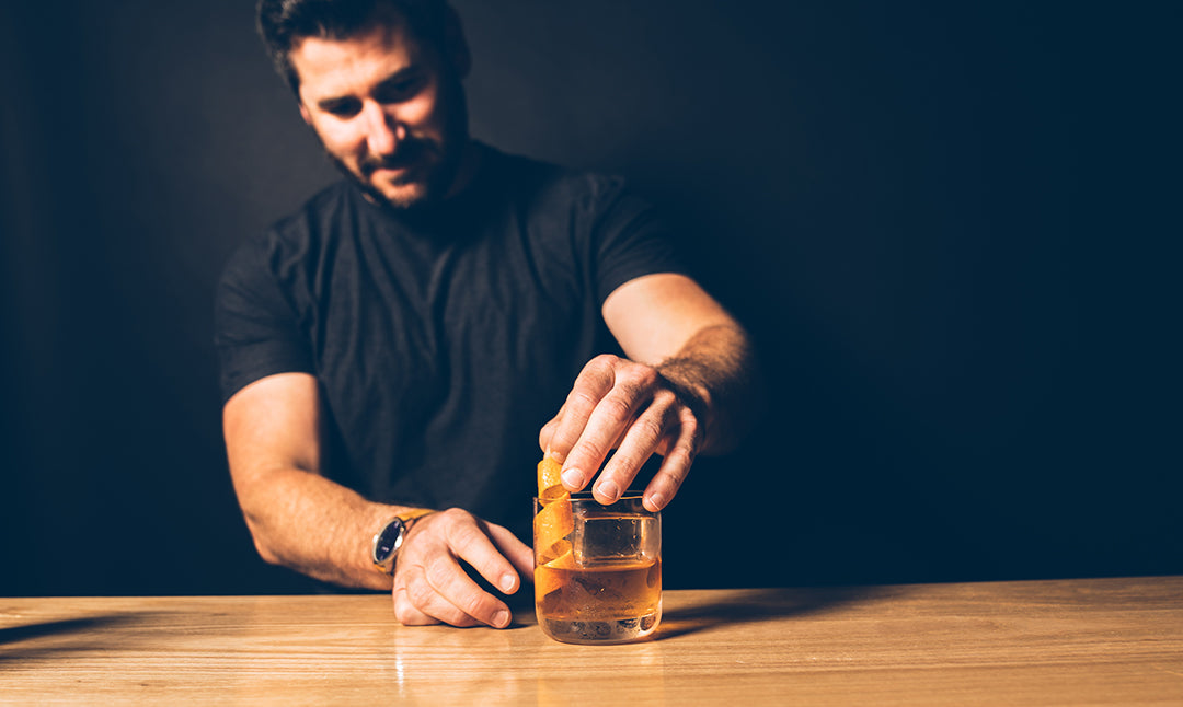 a man putting an orange twist garnish into a Sons & Dotters Old Fashioned cocktail with a large ice cube