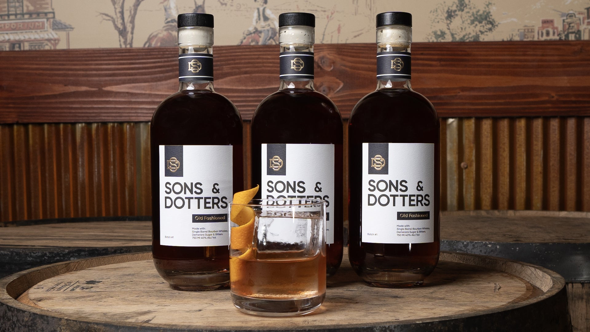 three bottles of Sons & Dotters with an Old Fashioned poured in a glass with an orange garnish and large ice cube on top of 3 bourbon barrels