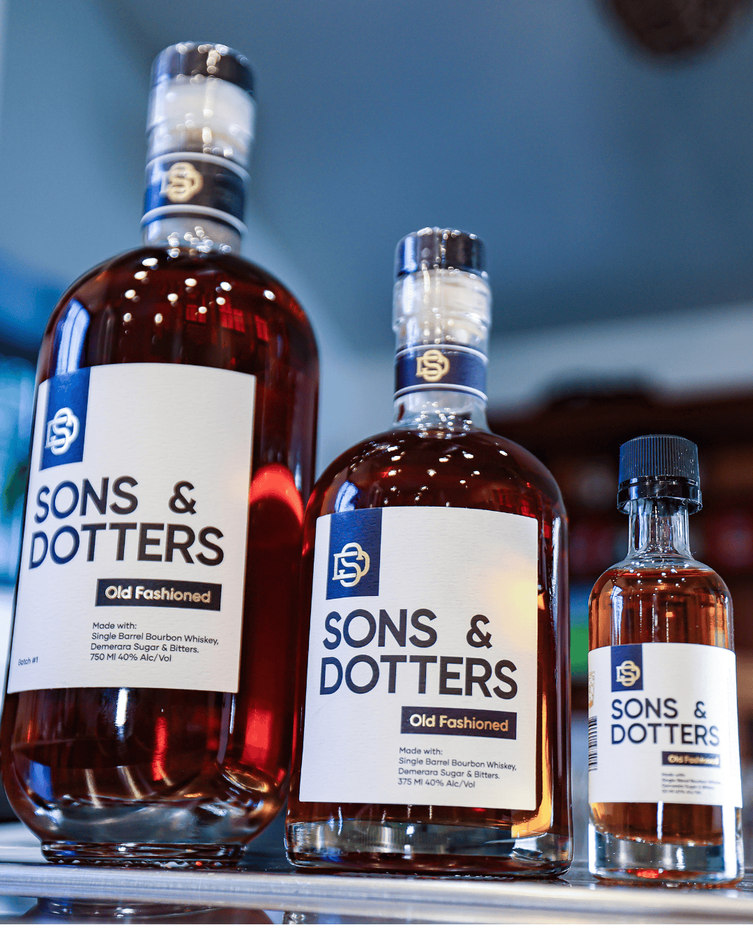 Three different sized bottles of Sons & Dotters Old Fashioned whiskey on a shelf.