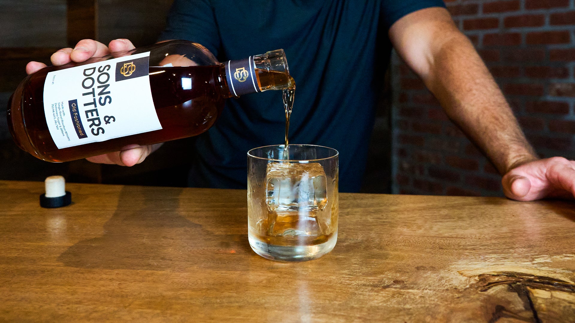 a bottle of Sons & Dotters Old Fashioned being poured out of the bottle into a glass with a large cube of ice and a cork resting on the bar