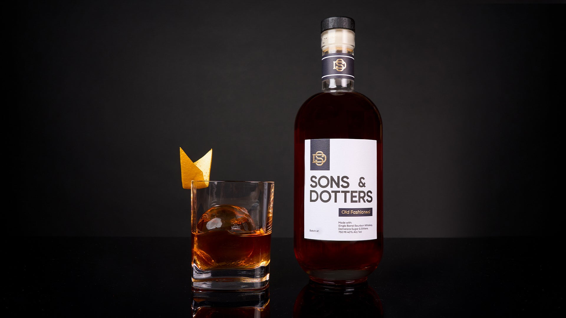 a prepared Sons & Dotters old fashioned in a rocks glass with a sphere of ice and a sharp orange garnish next to a bottle of Sons & Dotters Old Fashioned