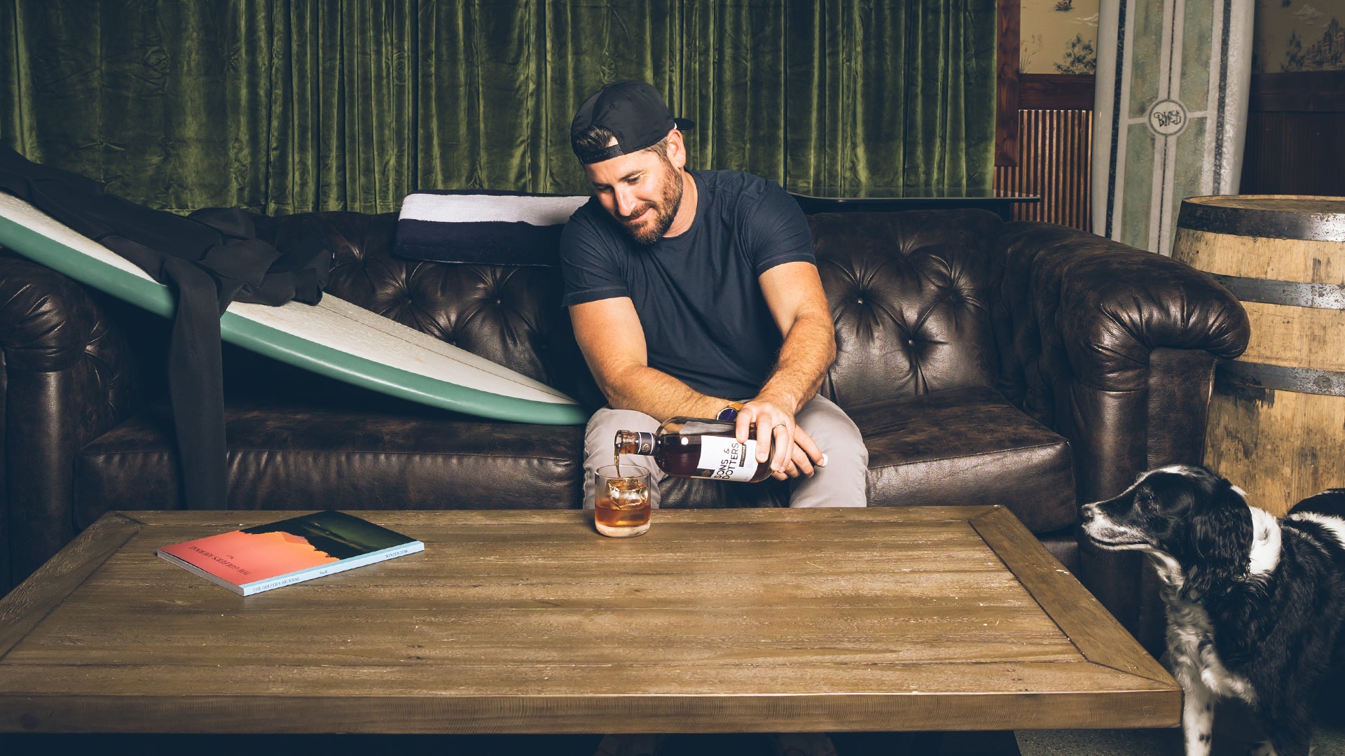 a man is sitting on the couch with a surfboard and wetsuit next to him as he pours a Sons & Dotters Old Fashioned on the table (with his dog next to the table0