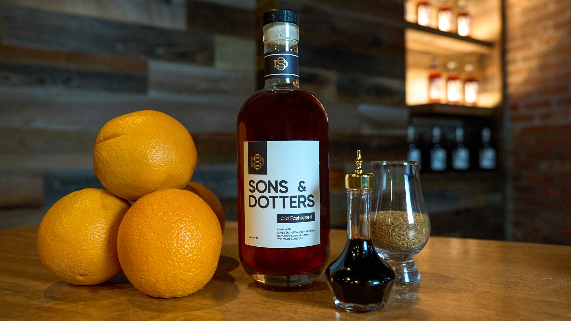 a bottle of Sons & Dotters Old Fashioned on a bar top with a bottle of bitters, a glass of demerara sugar and 4 oranges stacked next to it