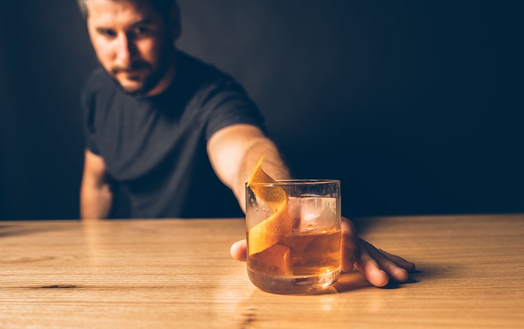 man behind the bar slides an old fashioned cocktail in a glass with an orange garnish across the bar